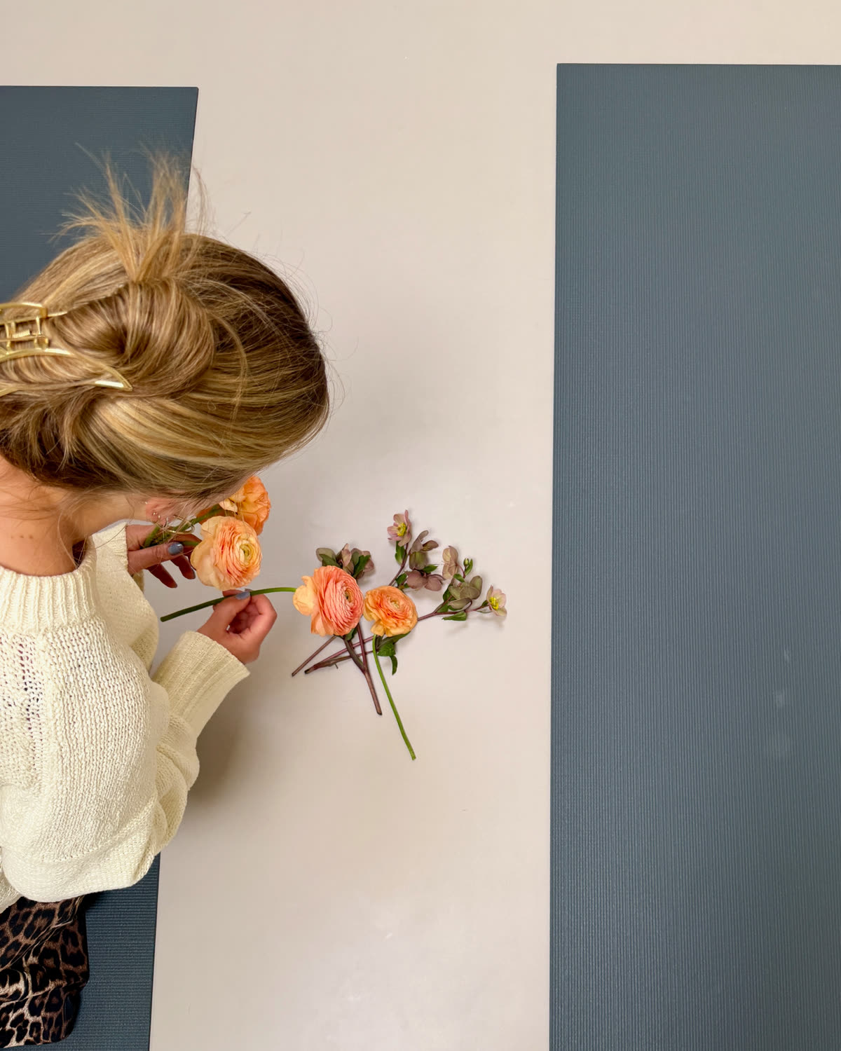 Top-down view of hands arranging peach ranunculus and small blossoms on a yoga mat