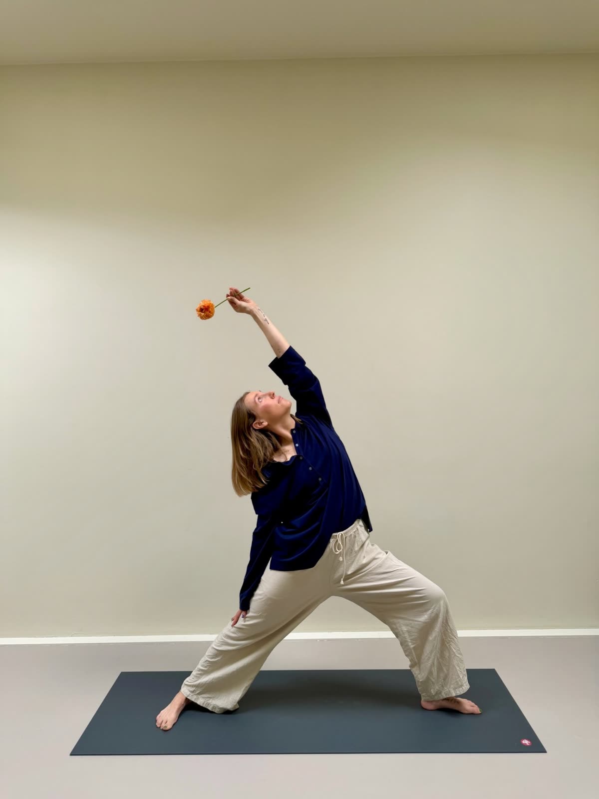 Kat in a warrior pose on a yoga mat, reaching one arm overhead with a single orange ranunculus